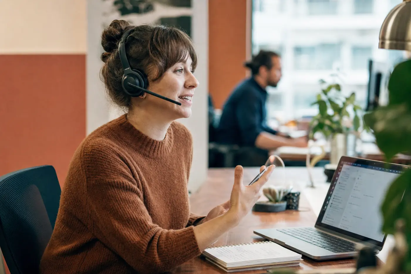 A customer success specialist mid-conversation on a headset, handwritten notes beside the laptop