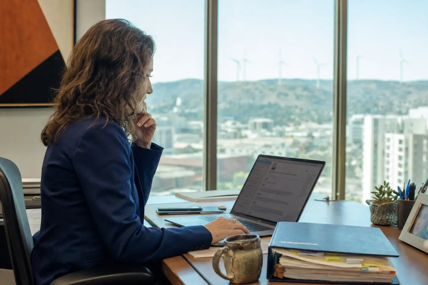 An energy lawyer searching a digital library of regulatory handbooks on a laptop, wind turbines visible through the office window